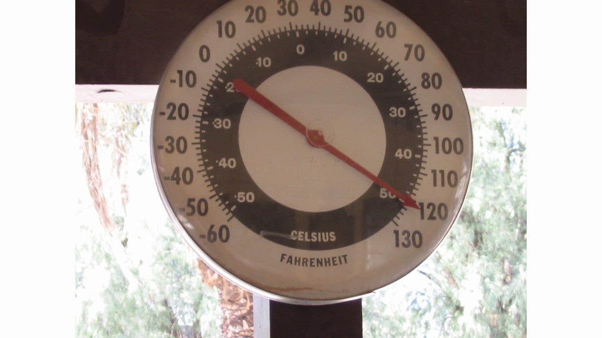 A large round dial thermometer with Celsius and Fahrenheit scales, its red needle pointing to about 120 degrees Fahrenheit, hanging in shade in Death Valley, California.