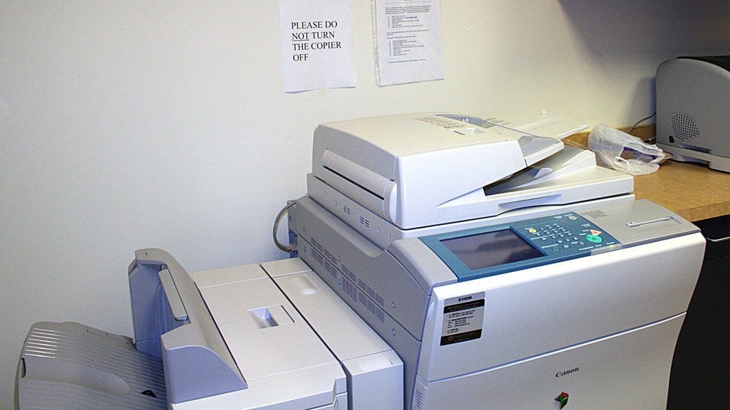 A large office multifunction copier and printer against a wall in an office workroom, with a printed notice above it reading please do not turn the copier off.