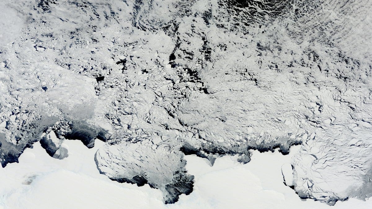 Satellite view of the East Antarctic coast, with the white ice sheet across the bottom and a broken belt of sea ice and icebergs drifting on dark ocean water above it.