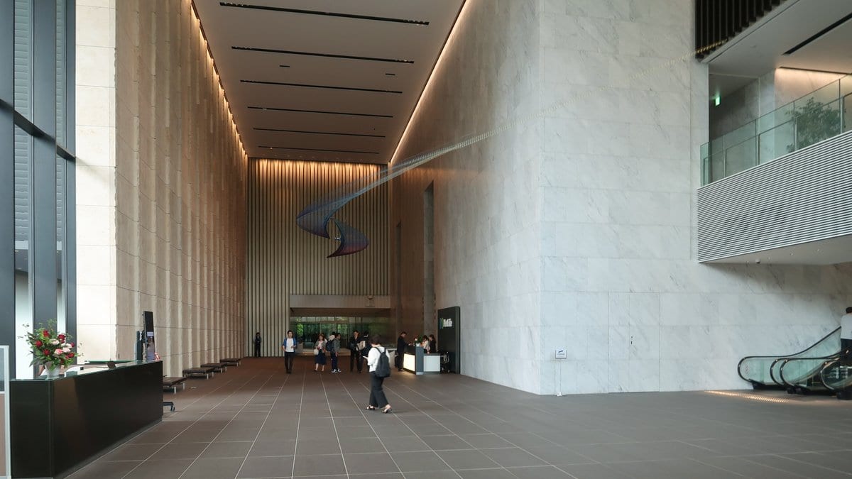 The tall, marble-lined lobby of a modern Tokyo office tower, with a reception desk at left and a few people walking toward the entrance.