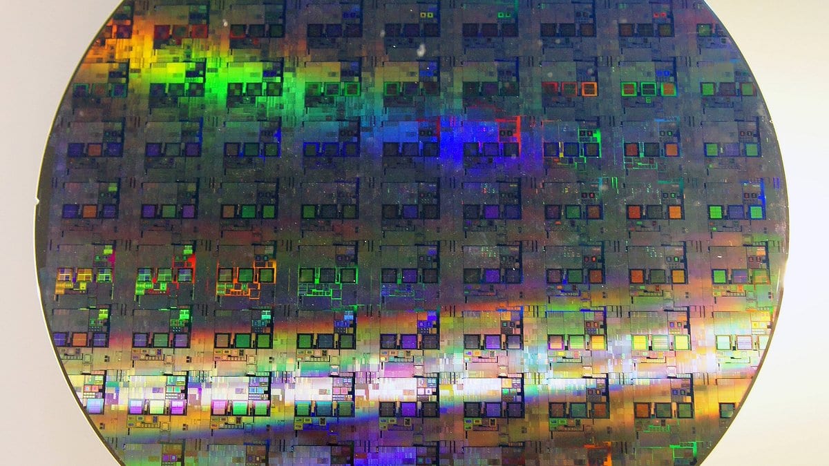 A circular silicon wafer covered in a grid of rectangular integrated circuit dies, diffracting light into green, blue and orange bands.
