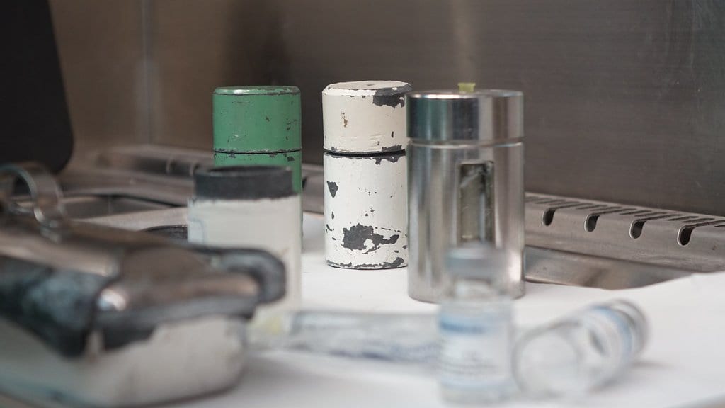 Lead shielding pots and small unlabeled glass vials on a white bench inside a stainless steel radiopharmacy hood.
