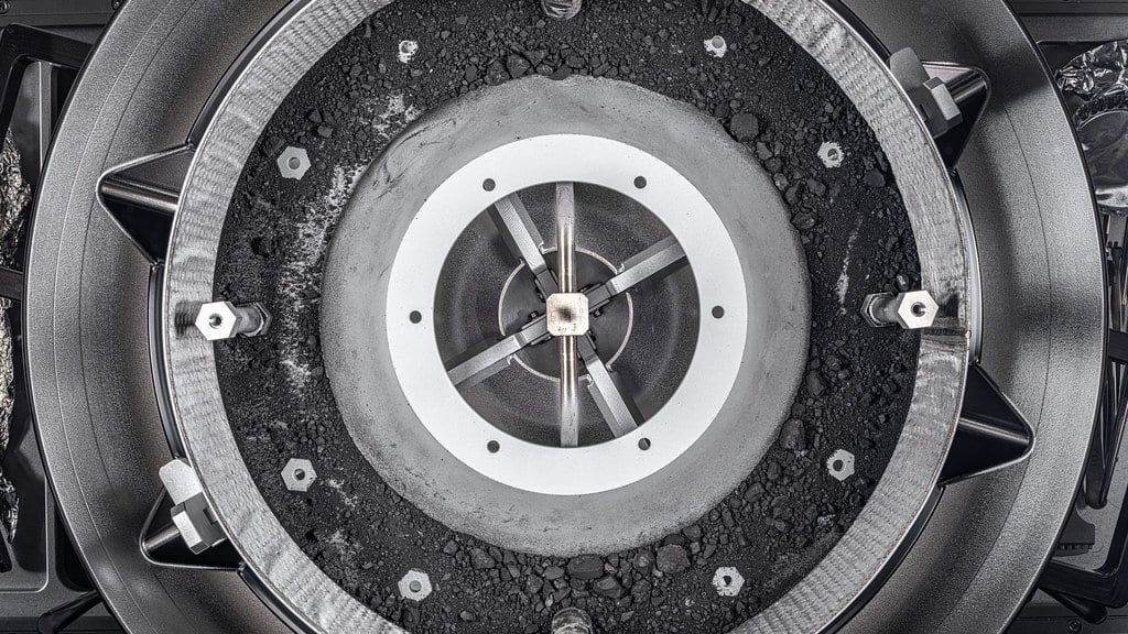 A circular sampler head seen from above, its inner ring packed with black asteroid dust and small rock fragments around a central collector.