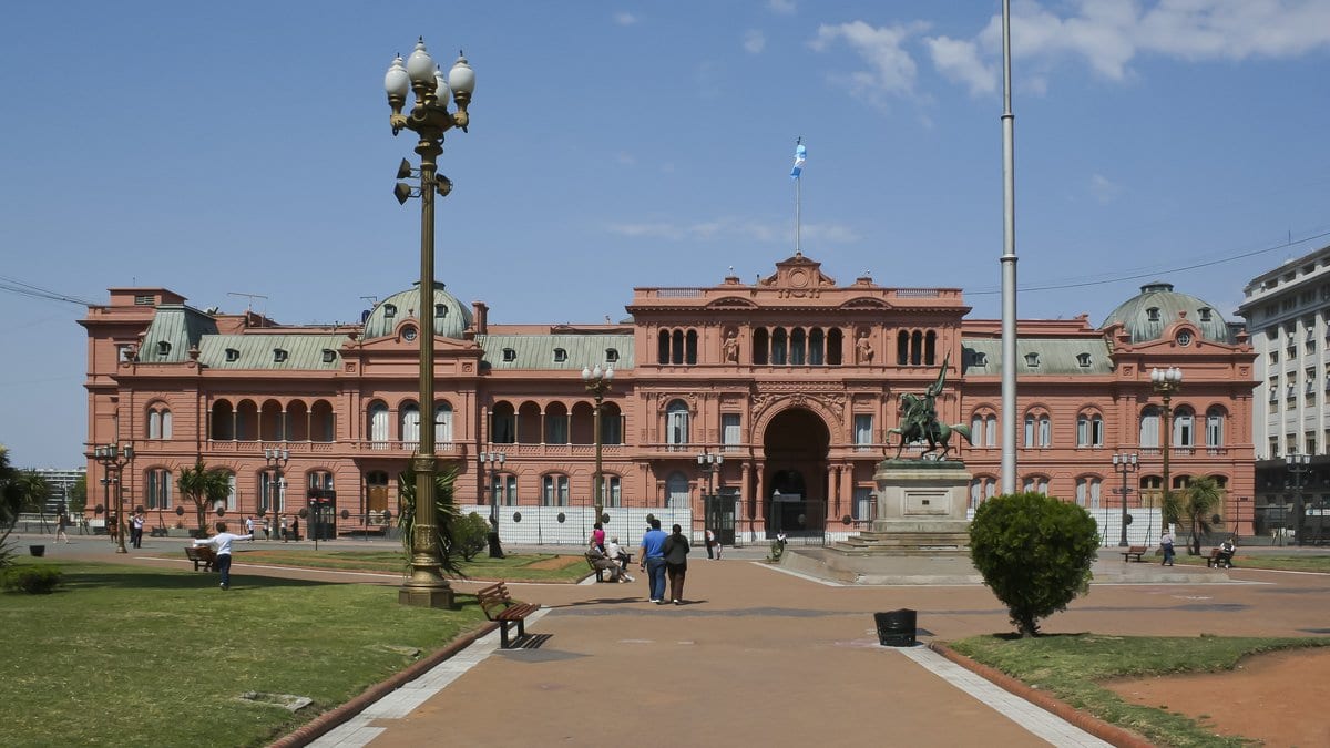 The pink neoclassical facade of the Casa Rosada presidential palace in Buenos Aires, seen across an open plaza under a blue sky.