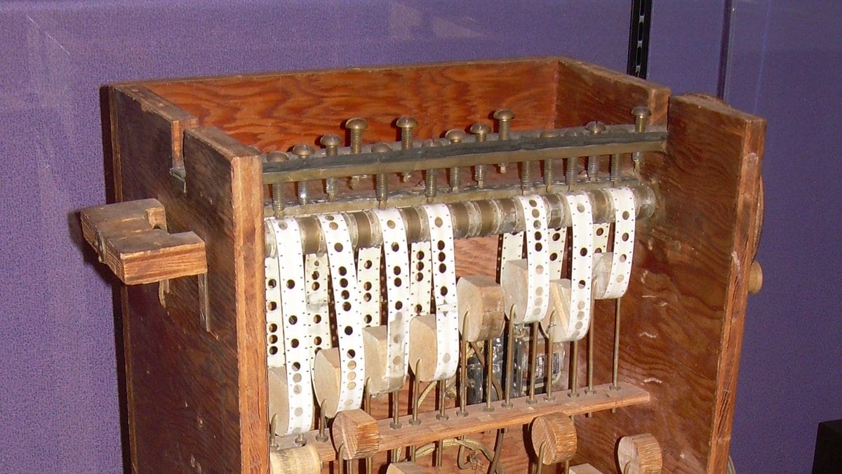 A wooden cabinet holding rows of punched steel chains and brass gears, an early machine for number theory calculations, inside a museum display case.