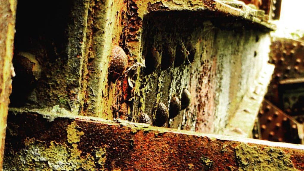 Close-up of a corroded riveted steel bridge joint, with rust swelling between the lapped plates and around the rivet heads.