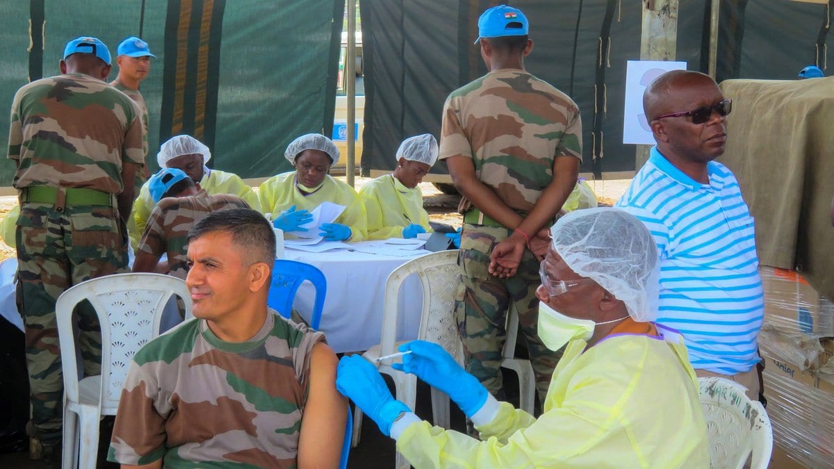 Health workers in yellow gowns, blue gloves and face masks give Ebola vaccinations to seated men at an outdoor vaccination point in Pinga, North Kivu.