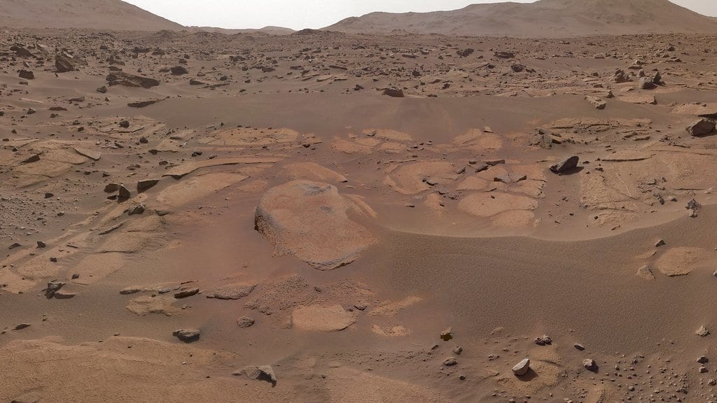 A rust-colored Martian plain of flat bedrock slabs, scattered dark rocks and rippled dust, with low hills along the horizon under a pale sky.