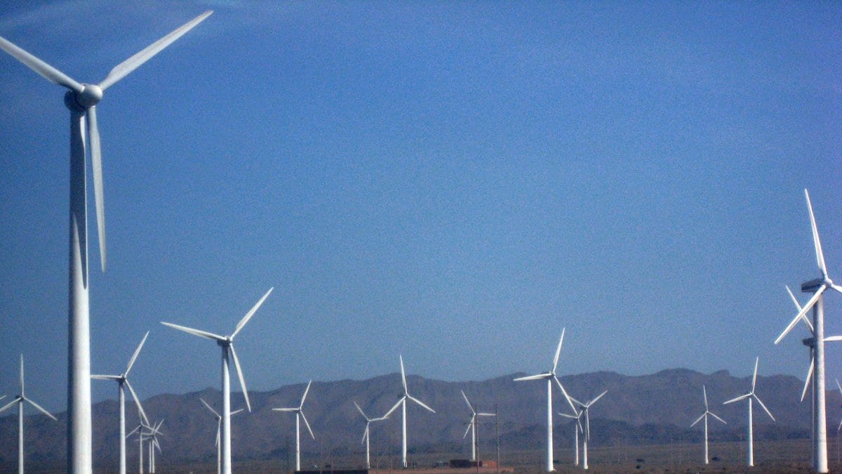 Rows of wind turbines on a gravel desert plain in Xinjiang, China, with a long mountain ridge behind them.