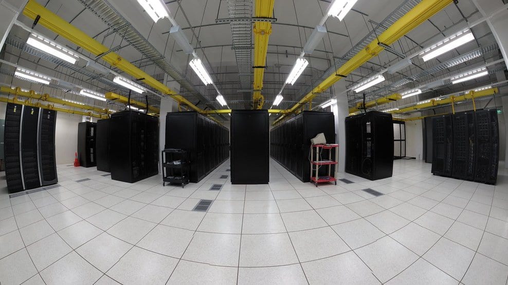 Rows of black equipment racks under yellow cable trays in a brightly lit data center hall with a white tiled floor.