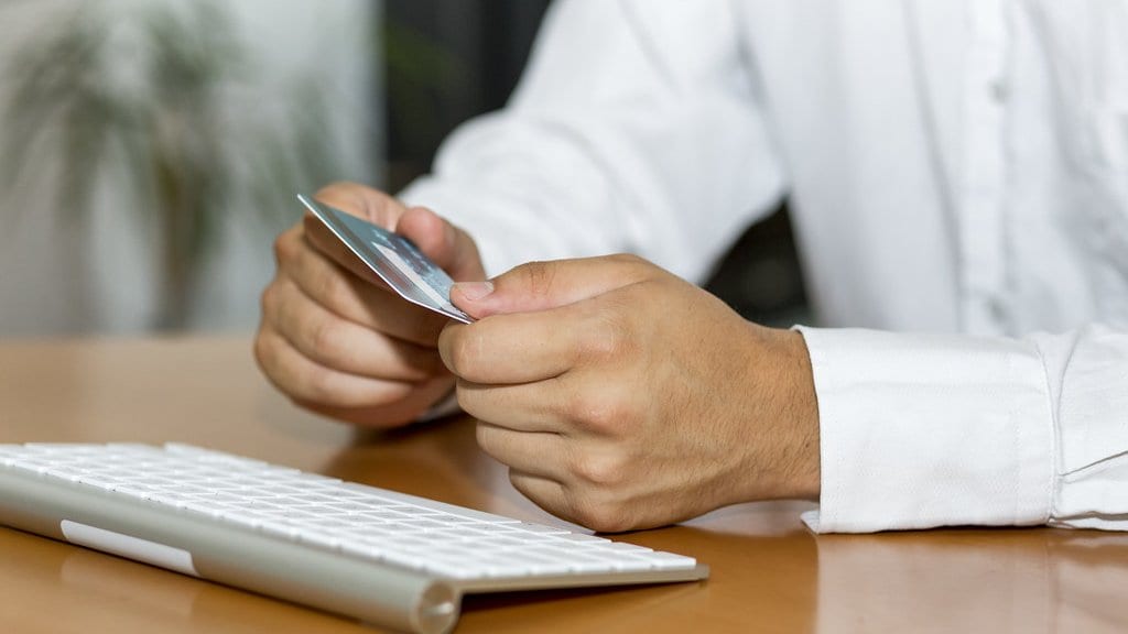 A person in a white shirt sits at a desk holding a payment card in both hands, with a computer keyboard in the foreground.