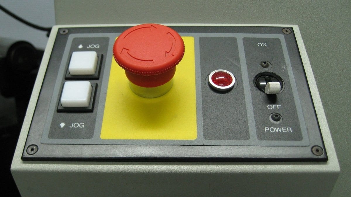 A red mushroom-shaped emergency stop button on a gray machine control panel, beside a toggle switch labeled ON and OFF.