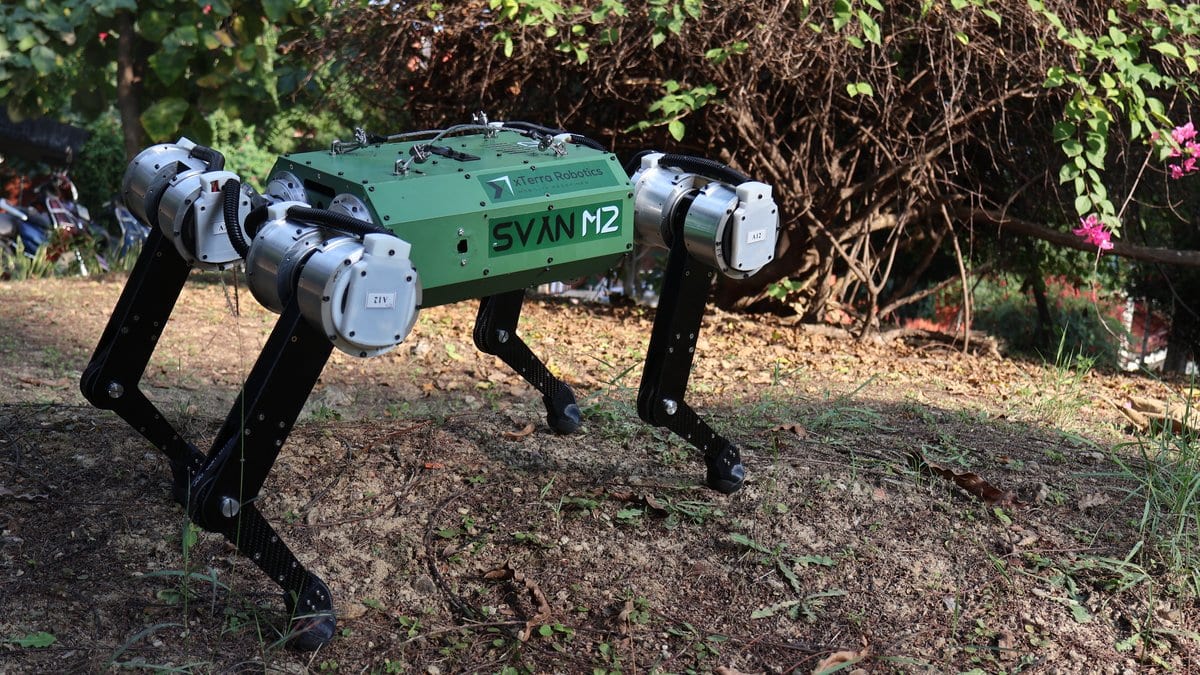 A green and black four-legged robot labeled SVAN M2 standing on rough ground in front of trees.