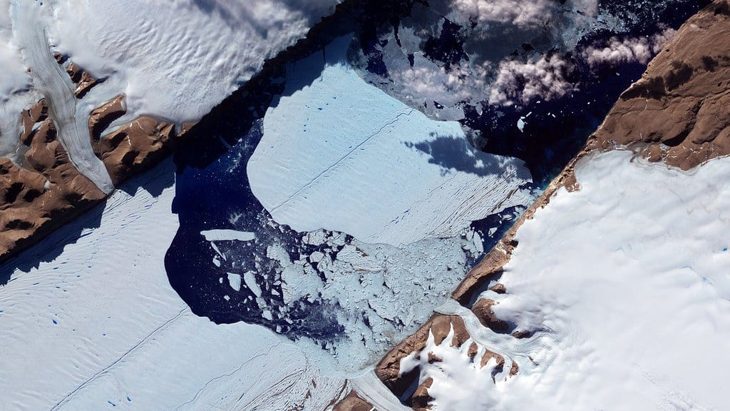 Satellite view of the ice front of Petermann Glacier in northern Greenland, with a large calved slab of ice surrounded by broken sea ice in the dark water of the fjord.