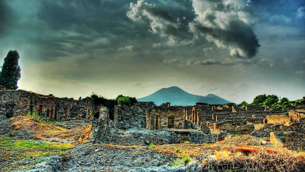 The excavated stone ruins of Pompeii in the foreground, with Mount Vesuvius rising behind them under a dark, clouded sky.