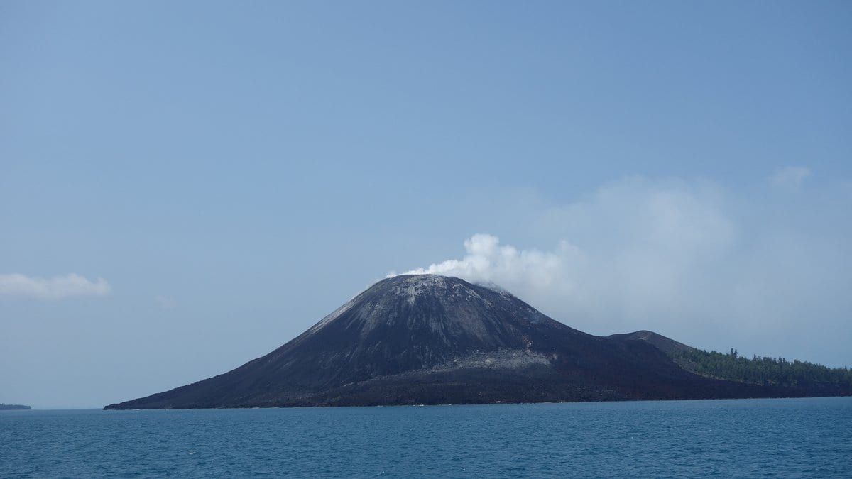 A dark volcanic cone on a small island rising from blue sea, with a white gas plume drifting from its summit.
