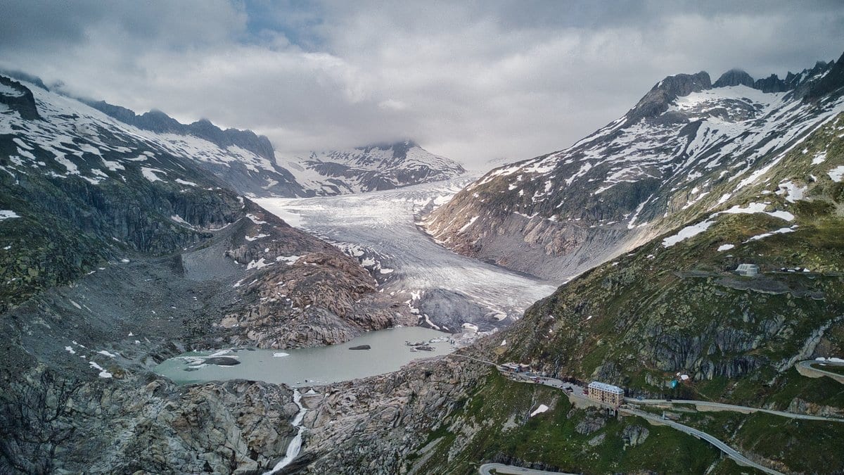 The tongue of the Rhône Glacier in the Swiss Alps ends in a gray meltwater lake ringed by bare rock, with a mountain road switchbacking up the slope at right.