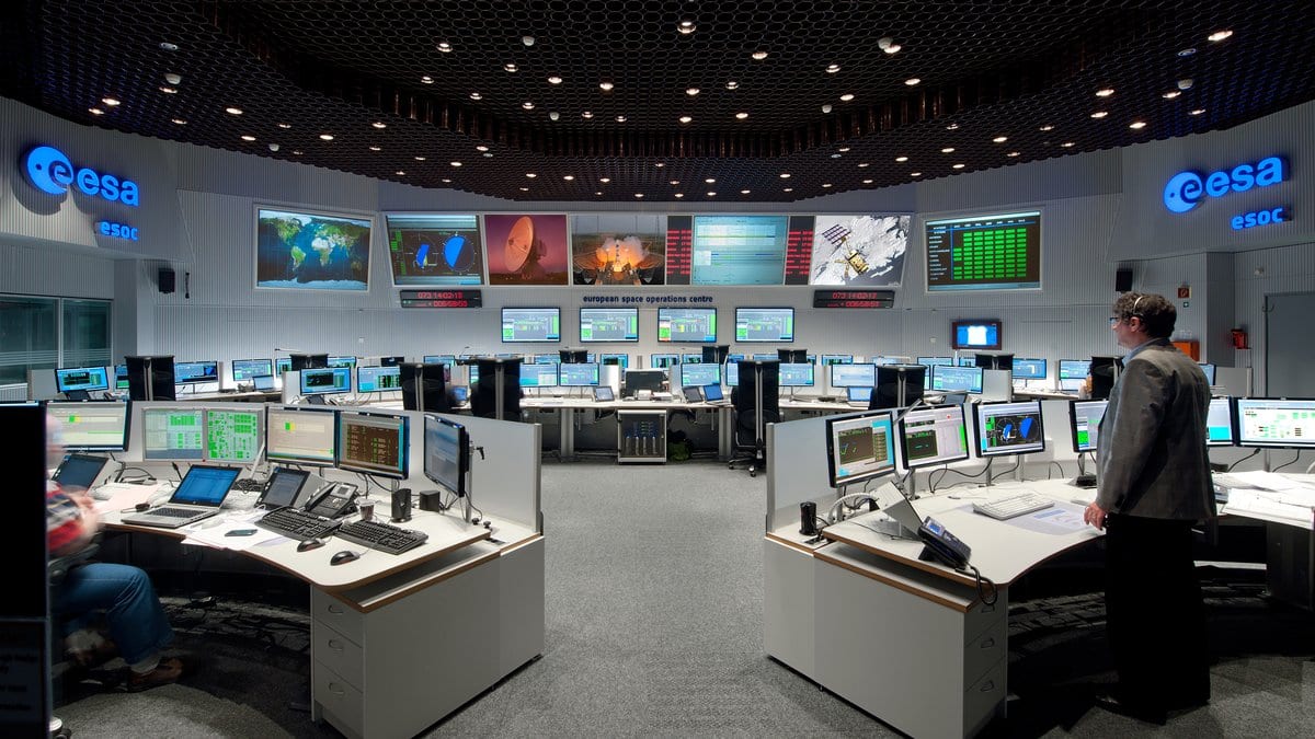 Operators at banks of consoles in a large spacecraft operations center, with world maps and telemetry displays on the far wall.