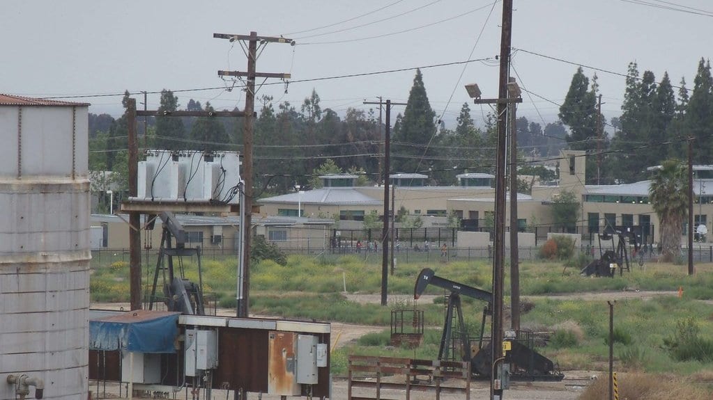 Two oil pumping units stand in a weedy lot behind a chain-link fence, with low school buildings and trees in the background.