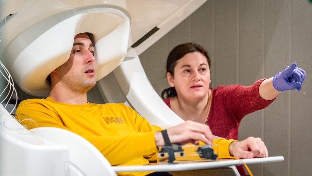 A young man reclines with his head inside the white helmet-shaped sensor array of a magnetoencephalography scanner while a researcher in a red sweater points to one side.