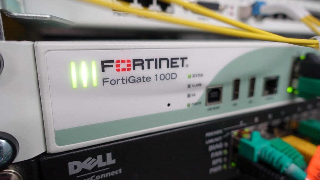 A white Fortinet FortiGate 100D network appliance mounted in a rack above a Dell PowerConnect switch, with yellow and green patch cables running to its ports.