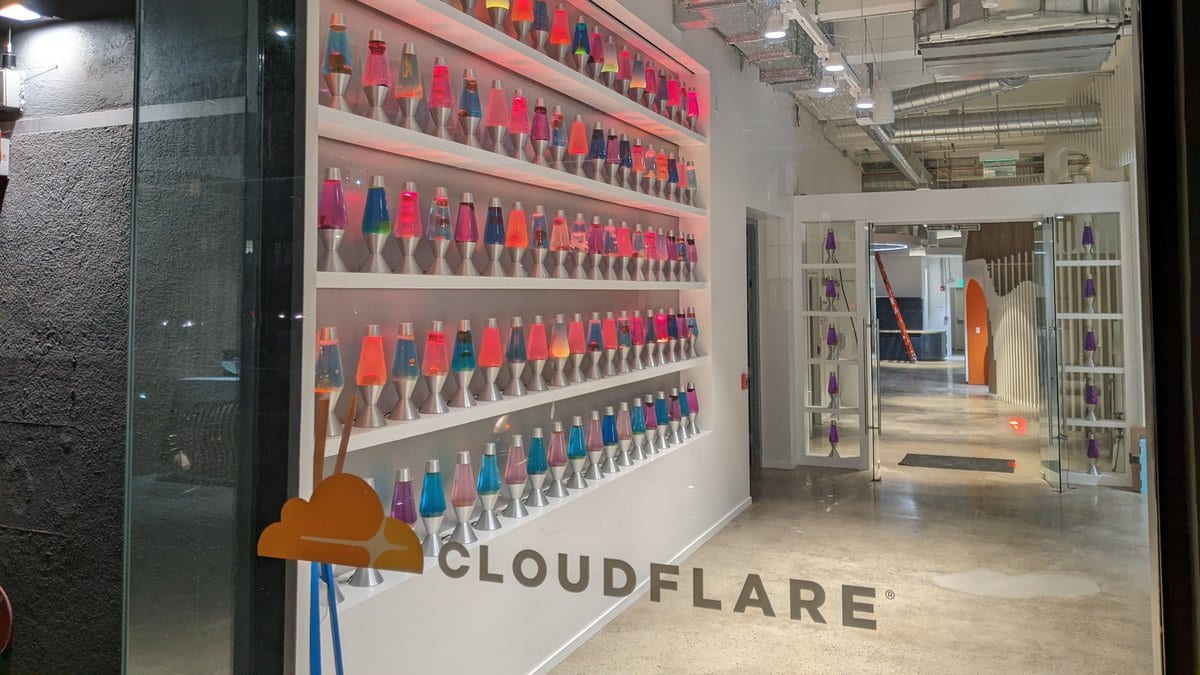 A wall of dozens of colorful lava lamps on white shelves beside a glass office entrance marked with the Cloudflare logo.
