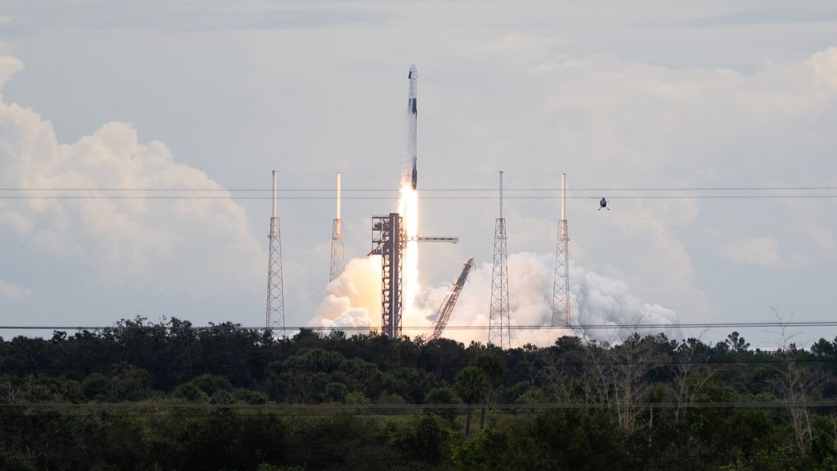 A SpaceX Falcon 9 rocket topped by a Dragon capsule rises past the lightning towers of its Florida launch pad, trailing flame and a wide white exhaust cloud.