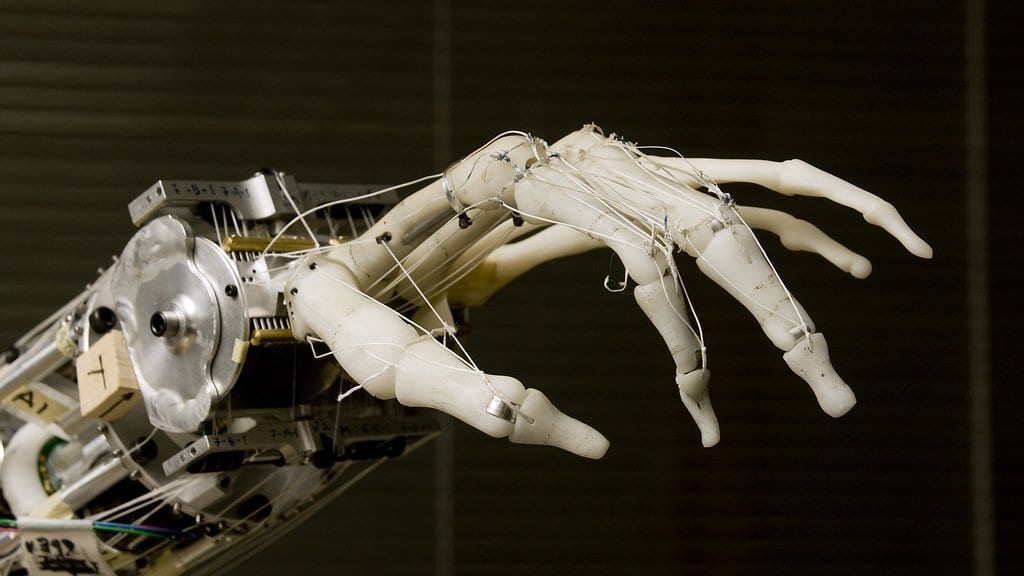 Close-up of a white anthropomorphic robot hand with jointed fingers pulled by thin cable tendons, mounted on a metal wrist in a laboratory.