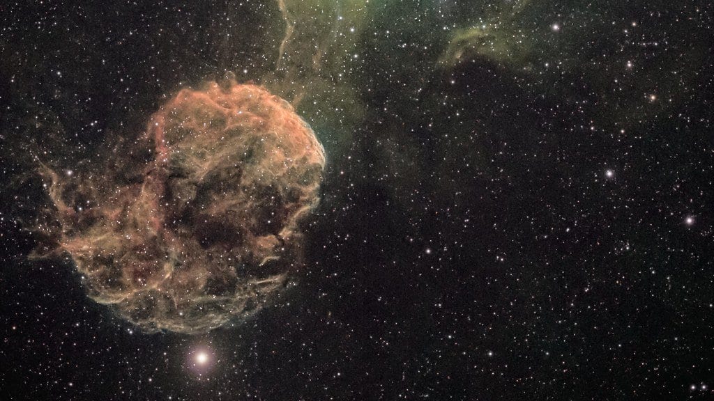 A rounded, filamentary supernova remnant shell glowing pink and orange against a dense field of stars, with faint green nebulosity above it.