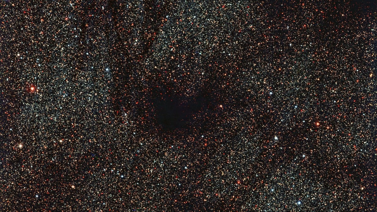 A dark patch of dust and gas blocks the light of a dense field of stars behind it.