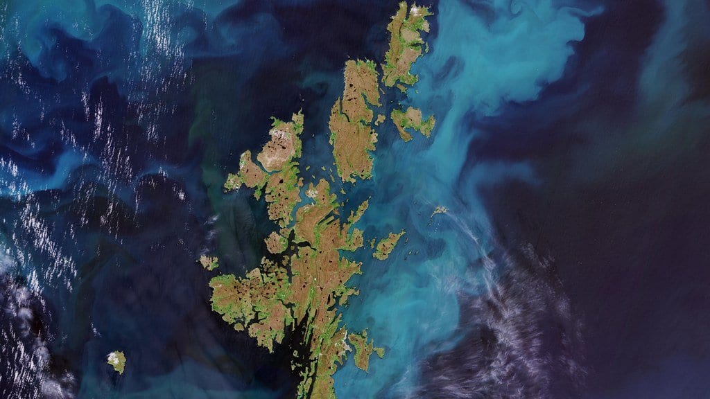 Satellite view of the Shetland Islands, brown and green, ringed by bright turquoise swirls of plankton in dark blue ocean water.