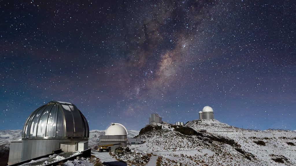 Observatory domes on a snow covered ridge at night beneath the band of the Milky Way.