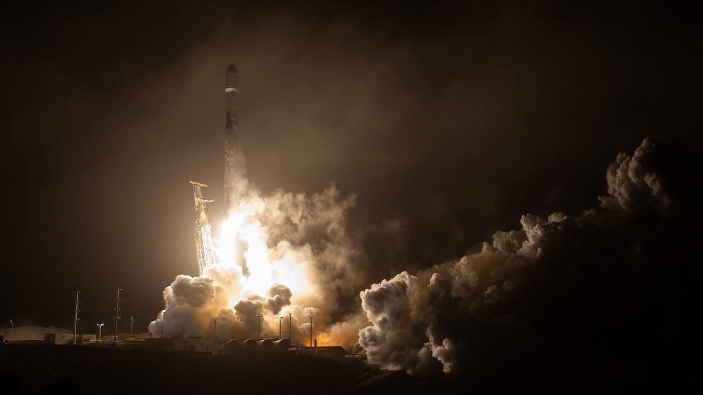 A Falcon 9 rocket lifts off at night, its engines lighting a wide cloud of exhaust across the launch pad.
