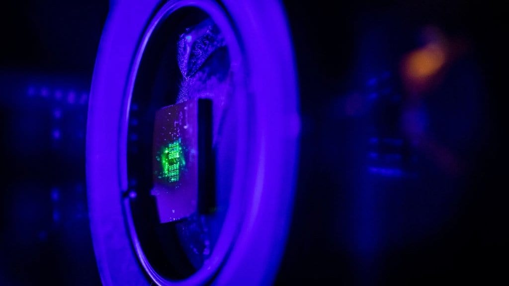 A small chip held in a round optical mount glows green where a laser strikes it, inside a darkened laboratory lit blue.