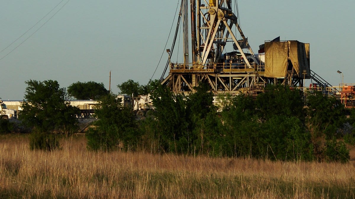 A steel drilling derrick and its rig floor rising above trees and dry grass at a natural gas well site, with service trucks parked alongside.