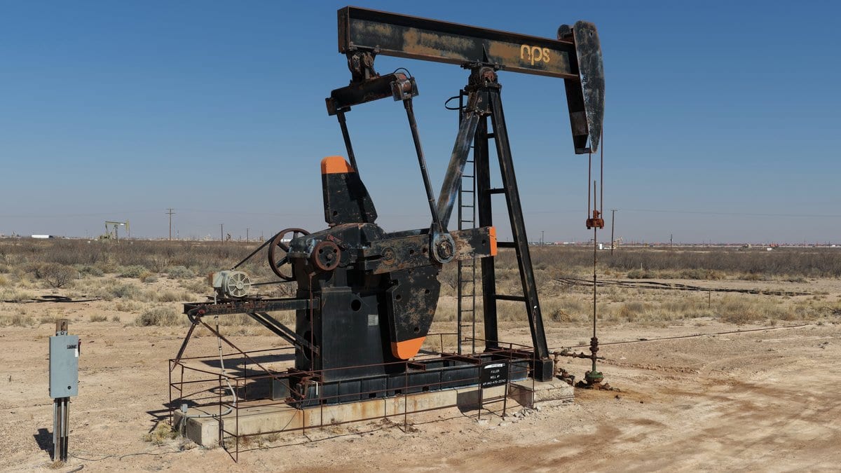 An oil pumpjack and wellhead equipment stand on a bare dirt well pad under a clear blue sky, with more production equipment along the flat horizon.