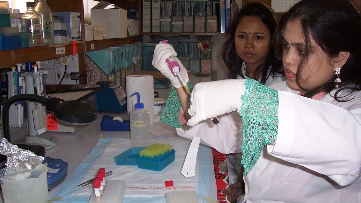 Two laboratory workers in white coats handle specimen tubes with a pipette at a bench in Bangladesh.