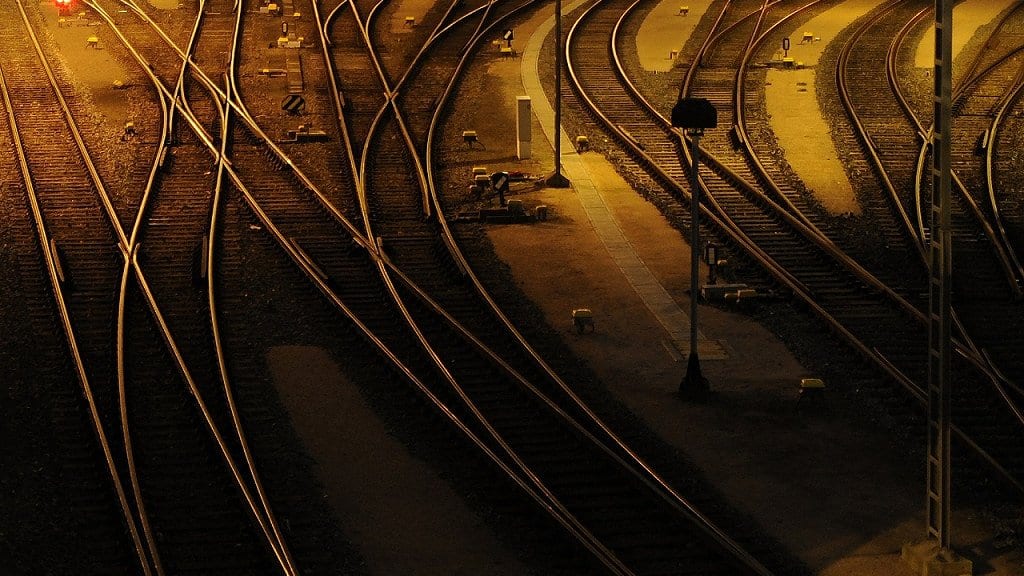 Many steel rail tracks crossing through switches in a rail yard at night, lit amber by overhead lamps.