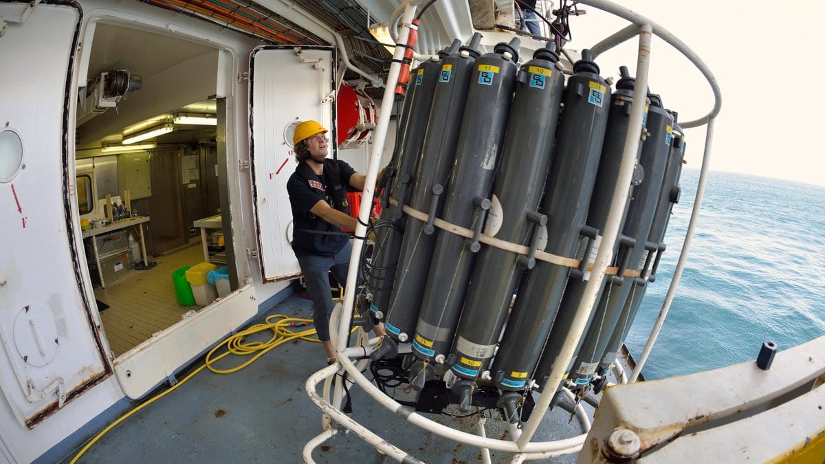 A crew member in a yellow hard hat guides a metal frame holding a ring of gray water sampling bottles aboard a research ship, with open ocean beyond the rail.