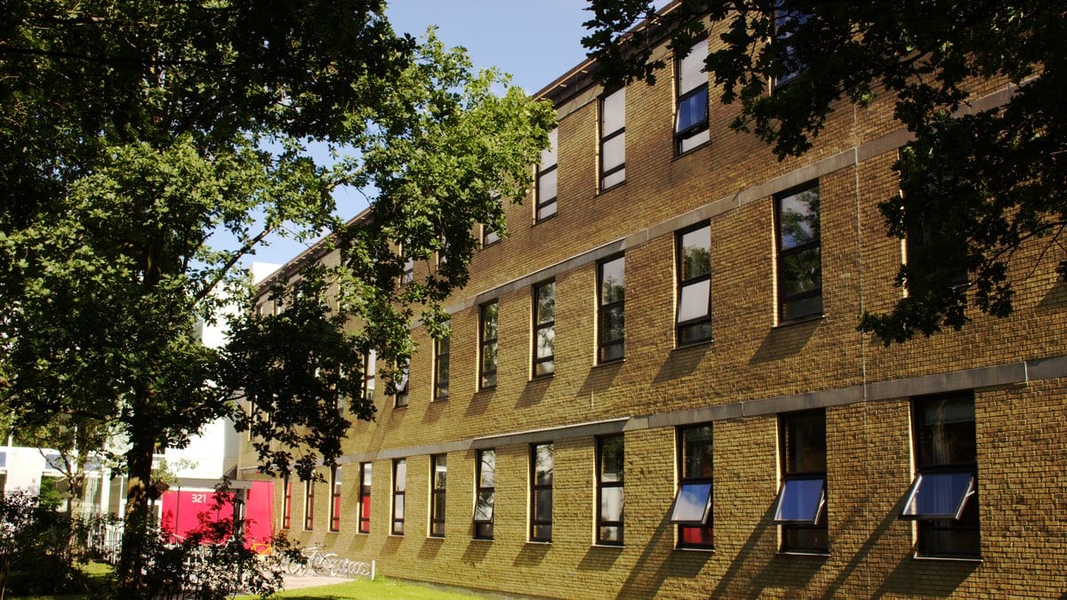 A yellow brick university building with rows of windows and awnings, framed by trees on a sunny day at the Technical University of Denmark.