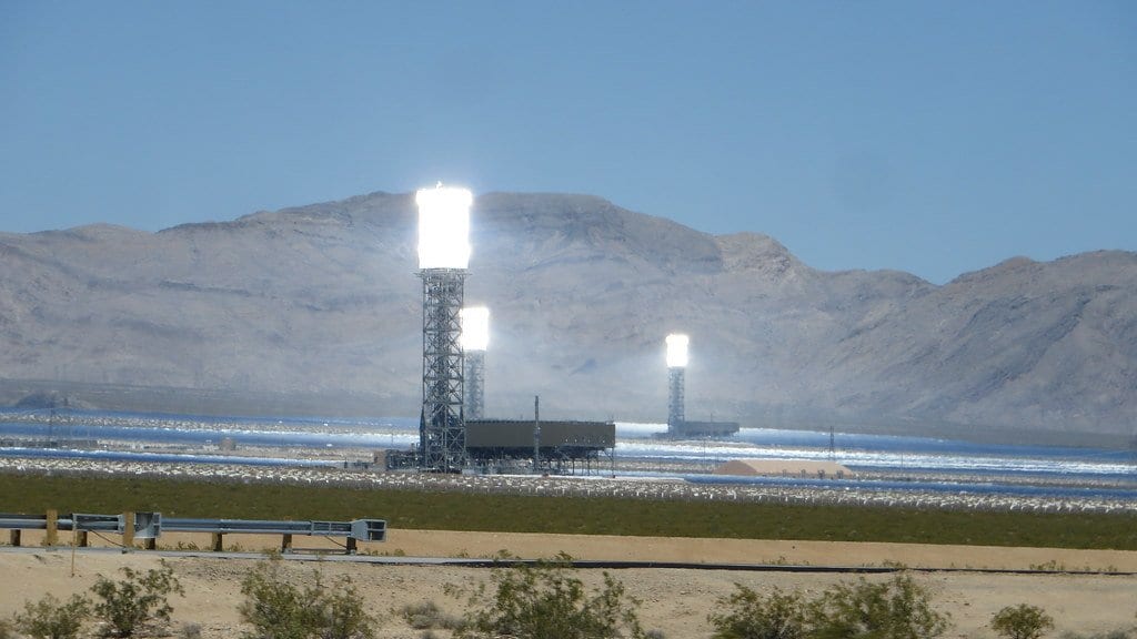 Three tall receiver towers glow white above a field of mirrors on a desert valley floor, with bare mountains behind.
