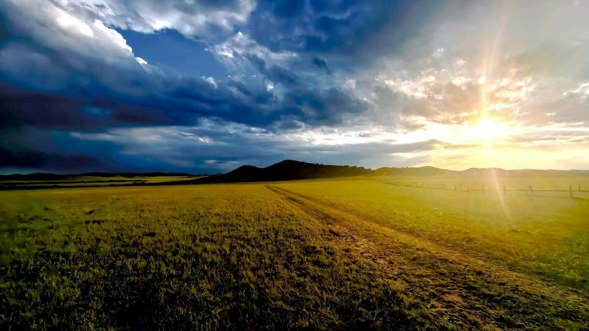 A dirt track crossing open grassland toward low hills, with the sun low on the horizon and heavy clouds above.
