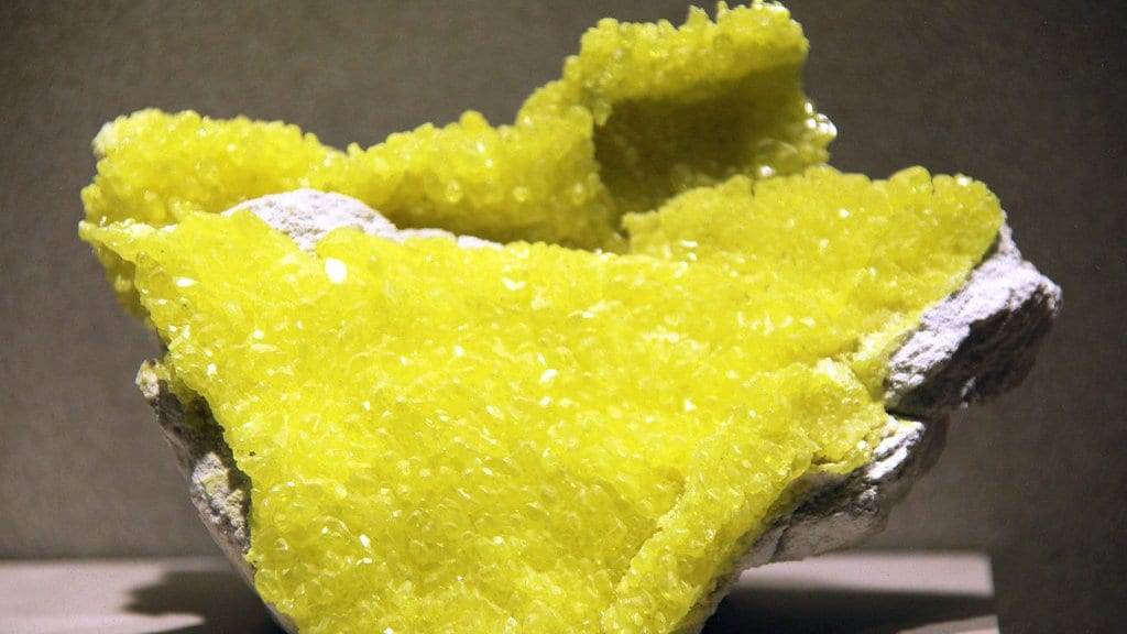 A bright yellow mass of native sulfur crystals on pale rock, shown on a museum stand.
