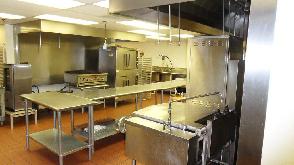 An empty stainless steel commercial kitchen, with a large extraction hood over a cooking range and prep tables, no people present.