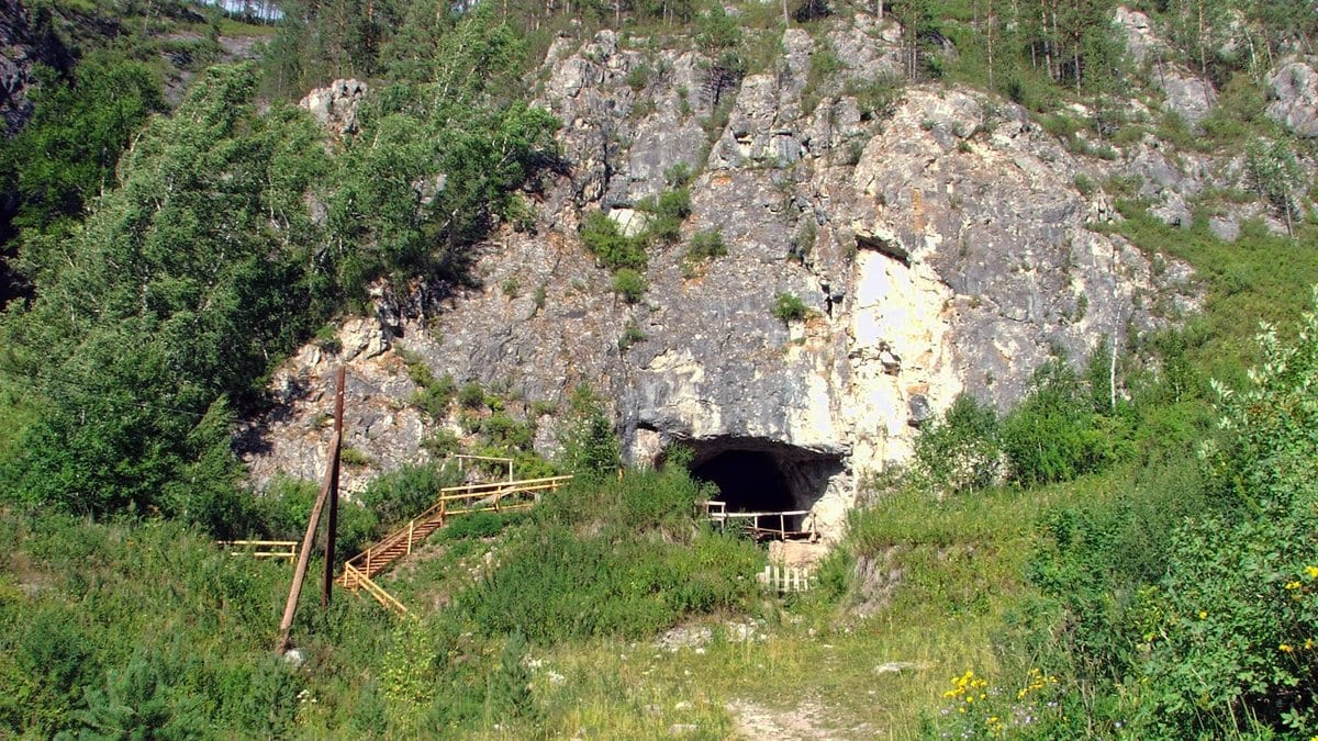 A cave entrance at the foot of a limestone outcrop on a forested hillside in the Altai Mountains, with a wooden stairway leading up to it.