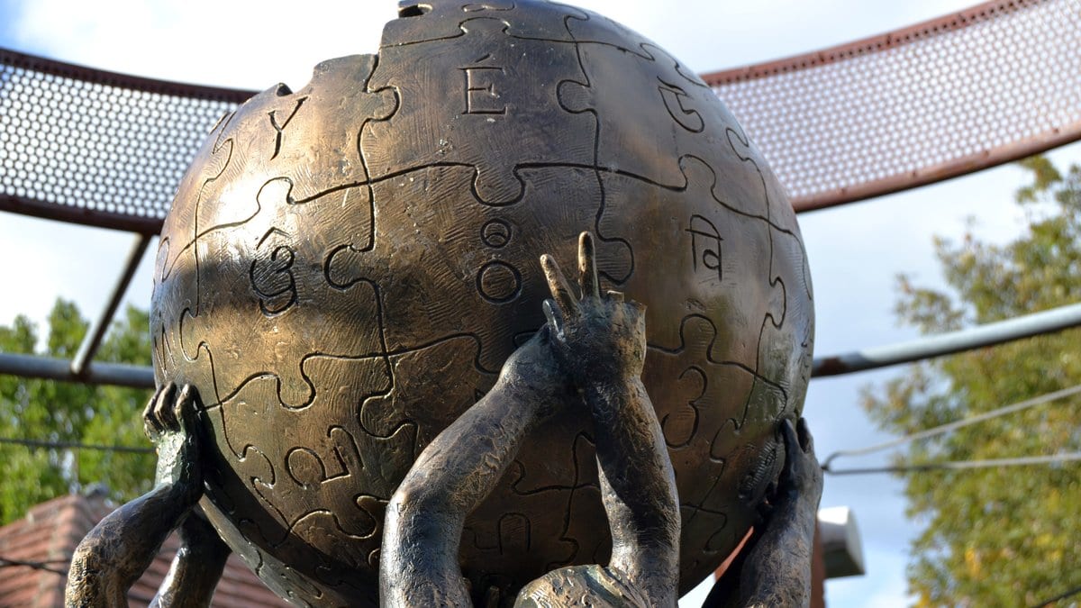 A bronze sculpture of human hands holding up a puzzle-piece globe against a cloudy sky.