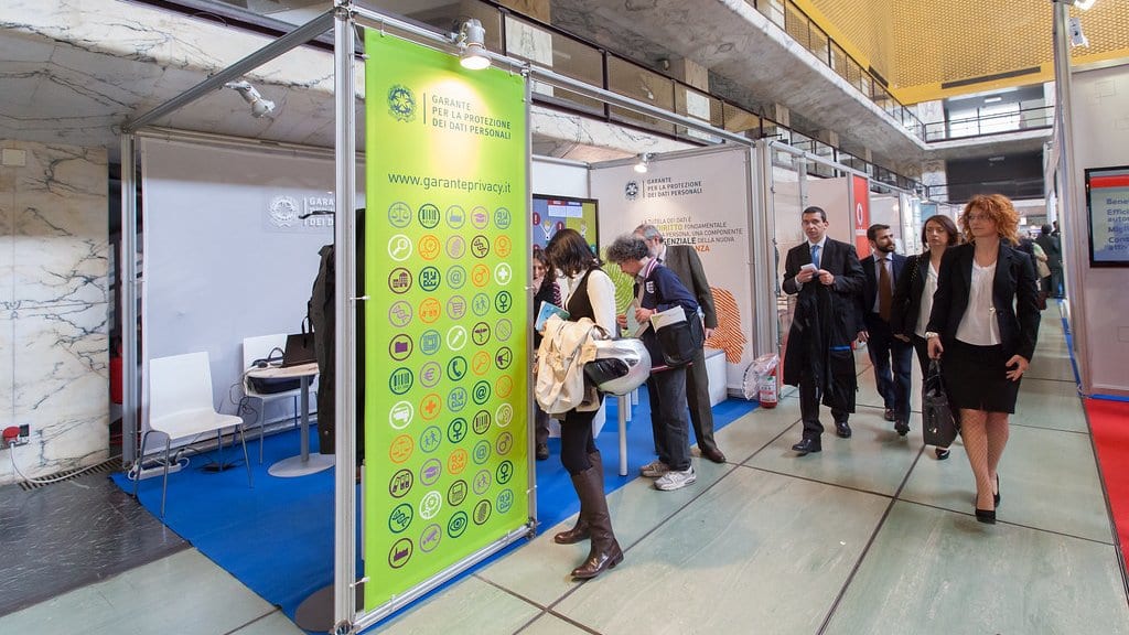 Visitors walk past the Italian data protection authority's exhibition stand, where a tall green banner carries the authority's name, logo and web address beside rows of small icons.