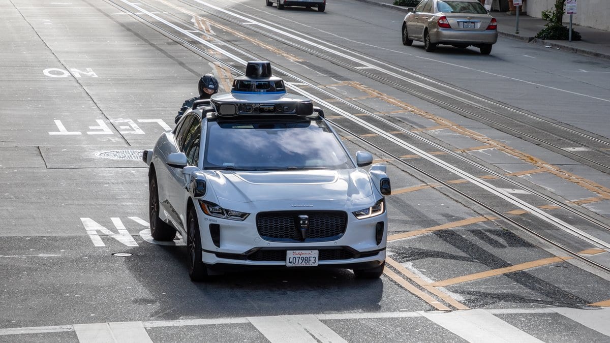 A white Waymo Jaguar I-Pace with roof-mounted laser scanners drives down a San Francisco street, a motorcyclist riding close behind it.