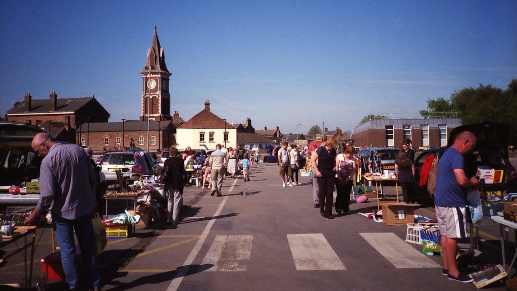 Shoppers and sellers along a row of stalls and open car boots piled with used goods at an outdoor market, a brick clock tower behind them.
