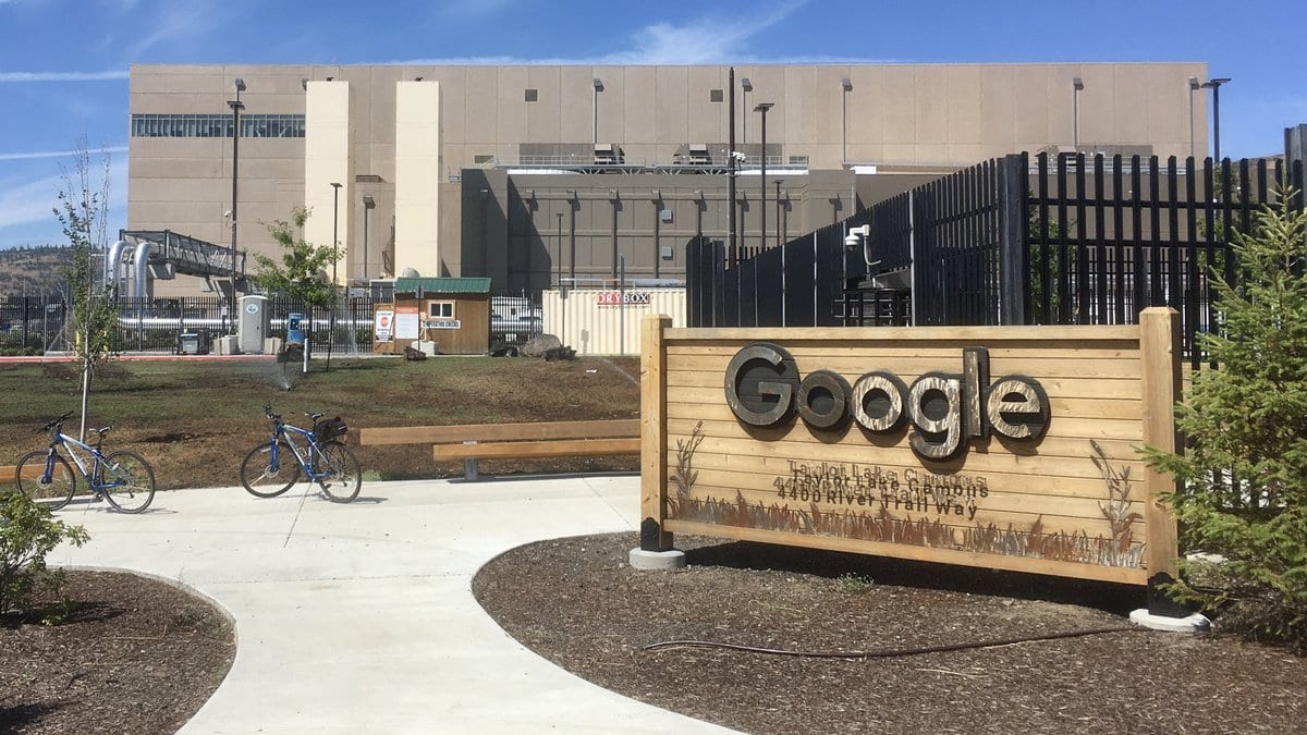 A long concrete data center building behind a security fence, with a wooden sign reading Google at the entrance path.